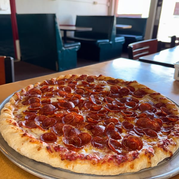 A large pepperoni pizza on a metal tray, with restaurant booths and tables blurred in the background.