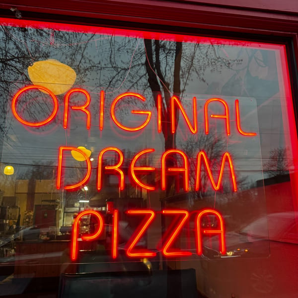 Red neon sign in a window reads "ORIGINAL DREAM PIZZA" with reflections of trees.