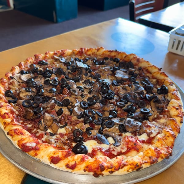 A whole pizza with black olives, mushrooms, and sun-dried tomatoes on a metal tray.