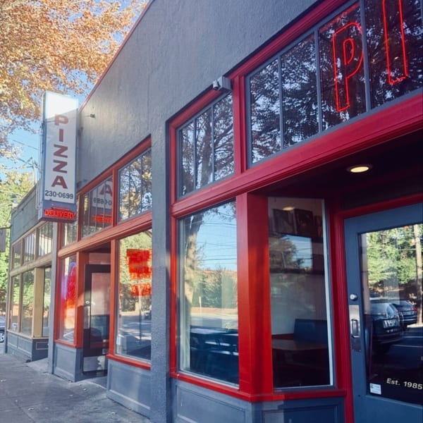 Exterior of a pizza restaurant with a vertical "PIZZA" sign and red window frames.