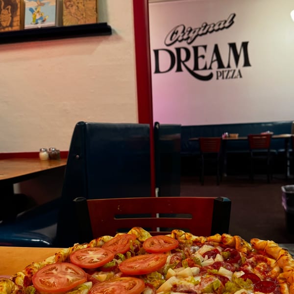A pizza with tomatoes, peppers, and pineapple sits on a table in a restaurant with a "Dream Pizza" sign.