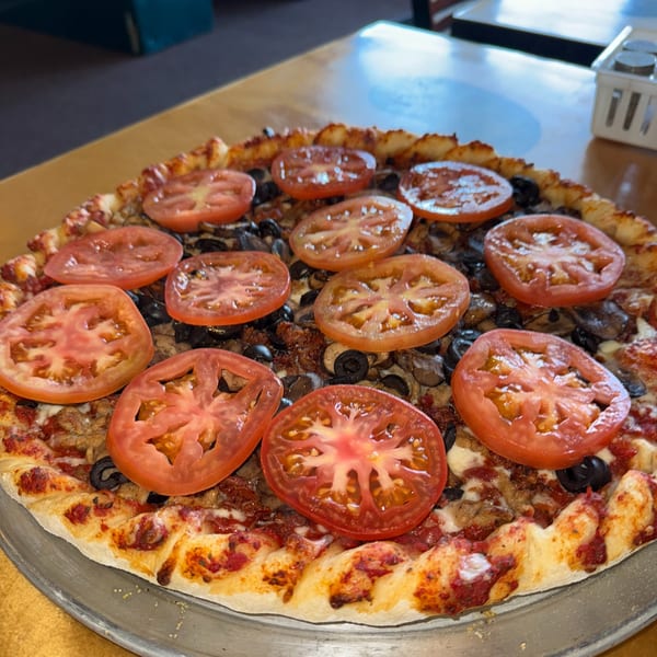 A whole pizza topped with sliced tomatoes, black olives, mushrooms, and sausage on a metal pan.
