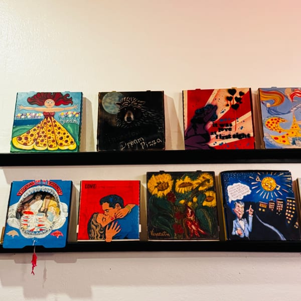A collection of colorful, whimsical pizza-themed artworks displayed on two black shelves against a white wall.