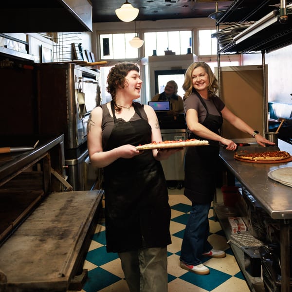 Two women in aprons preparing pizzas in a brightly lit commercial kitchen with checkered floors.