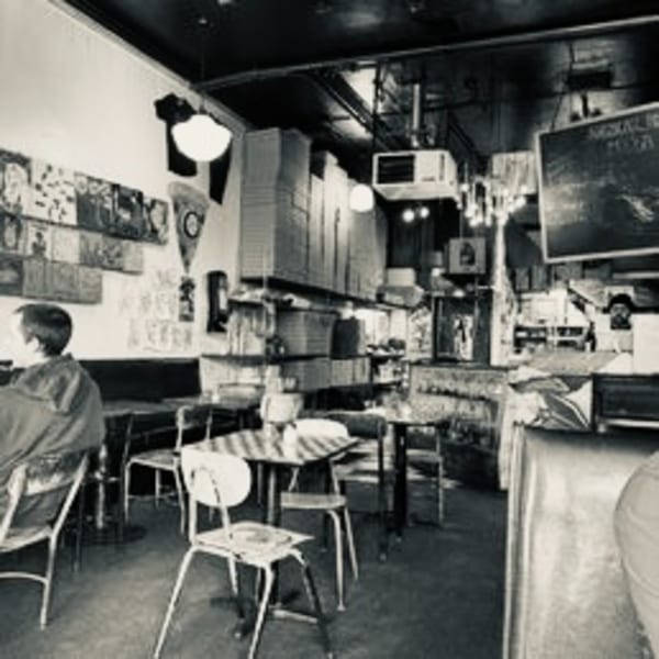 Black and white interior of a casual restaurant with a person seated, tables, chairs, and wall decor.