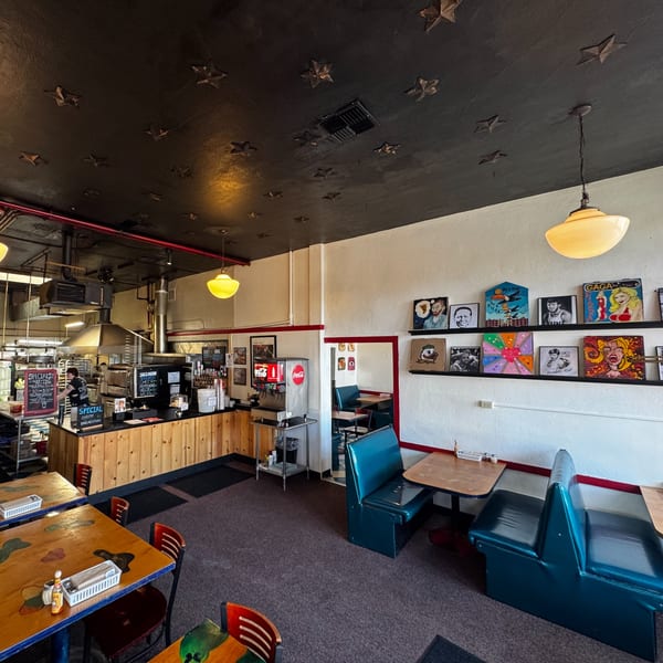 Interior of a casual restaurant with blue booths, wooden tables, and a counter with kitchen equipment.
