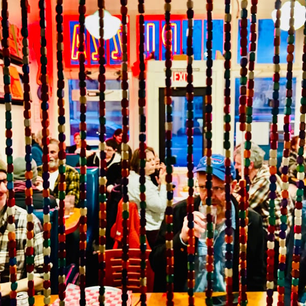 Diners seen through colorful beaded curtains, with a neon sign and exit sign in the background.